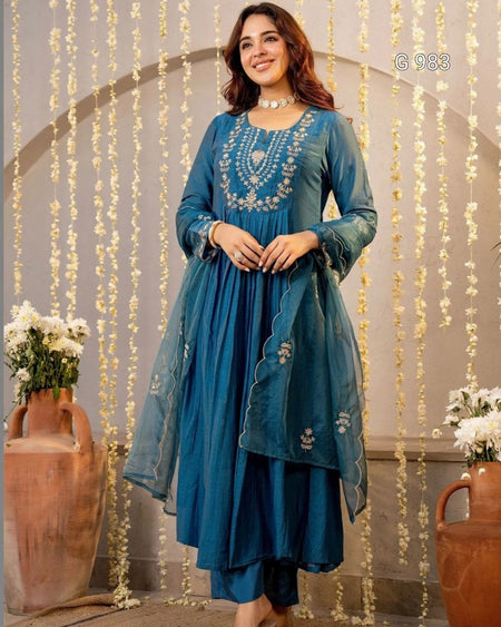 A woman models a teal blue ethnic salwar kameez suit with intricate silver embroidery on the chest, paired with a matching sheer dupatta, set against an arched backdrop with hanging floral garlands.
