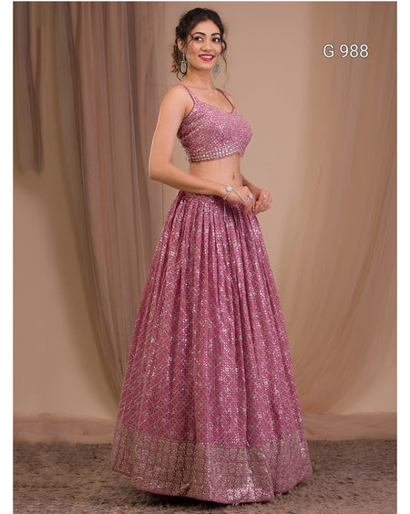 A woman modeling a pink heavily embellished Indian ethnic wear set consisting of a sleeveless crop top and a full flared skirt, with a beige draped backdrop.