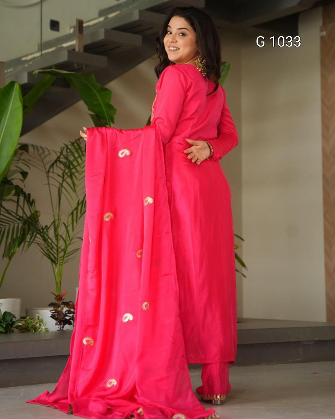 A woman modeling a bright pink traditional Indian outfit with a matching dupatta featuring small gold paisley motifs, standing indoors near plants and stairs. The text 'G 1033' appears in the top right.
