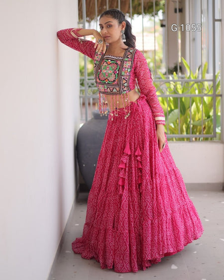 A woman models a bright pink traditional Indian lehenga choli set with intricate multicolor embroidery on the bodice, standing on an outdoor balcony.