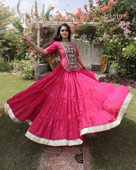 A woman outdoors in a garden wearing a bright pink traditional Anarkali gown with a flared skirt and embroidered neckline. The dress has cream border trim and a heavily embellished embroidered chest panel.