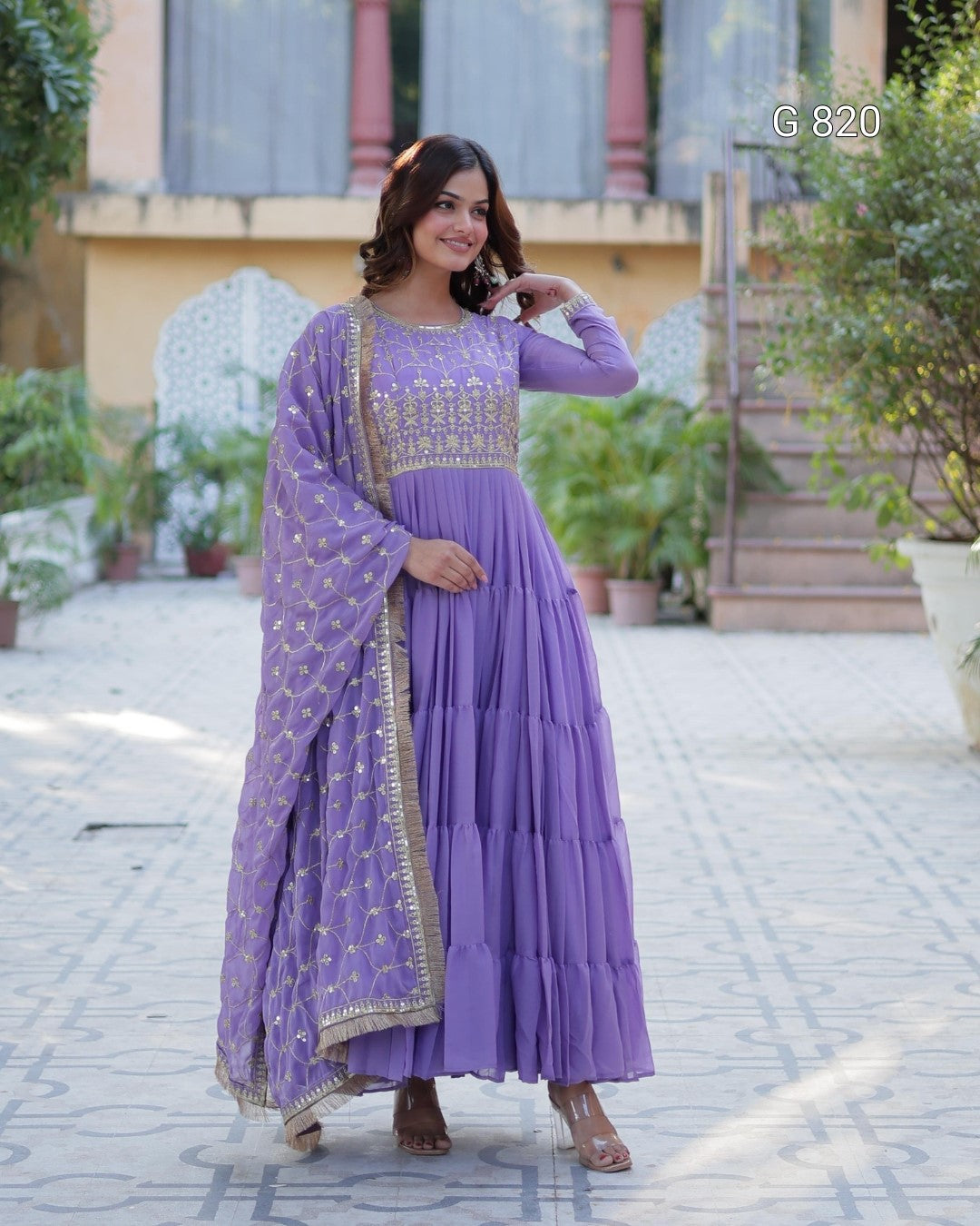 Woman in a purple traditional outfit standing outdoors with plants and a building in the background.