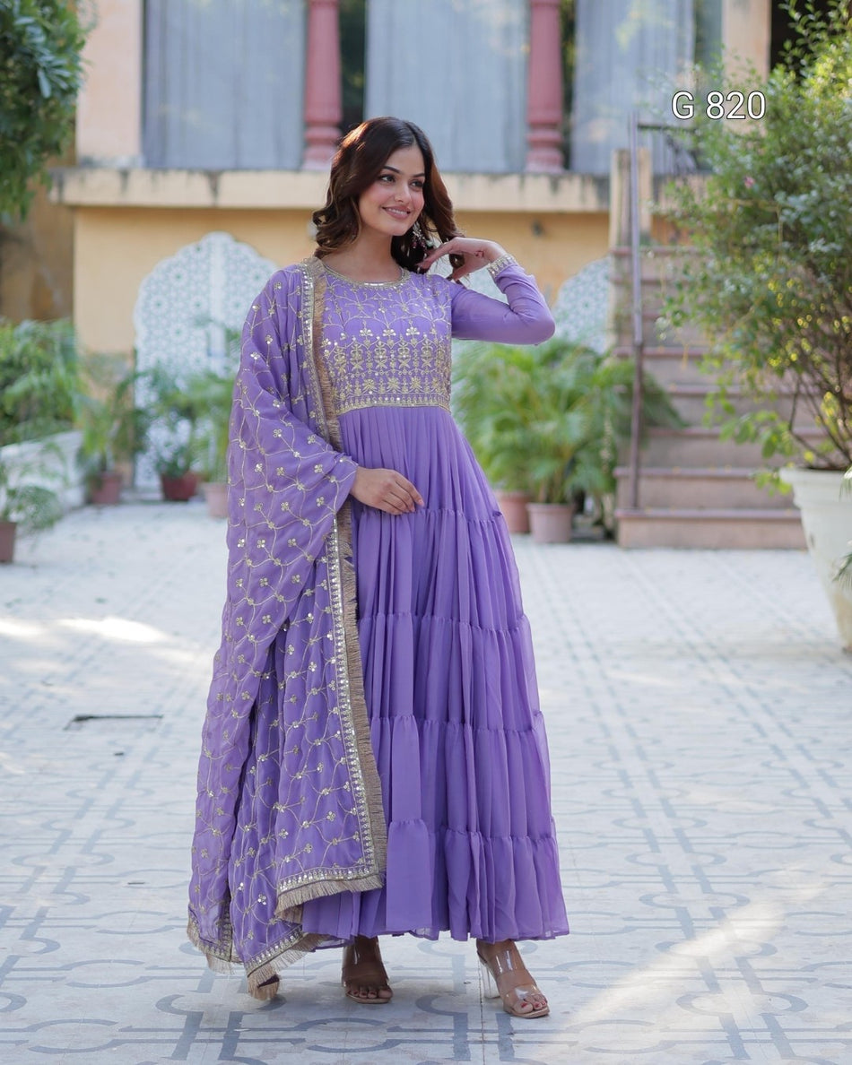 Woman in a purple traditional outfit standing outdoors with plants and a building in the background.