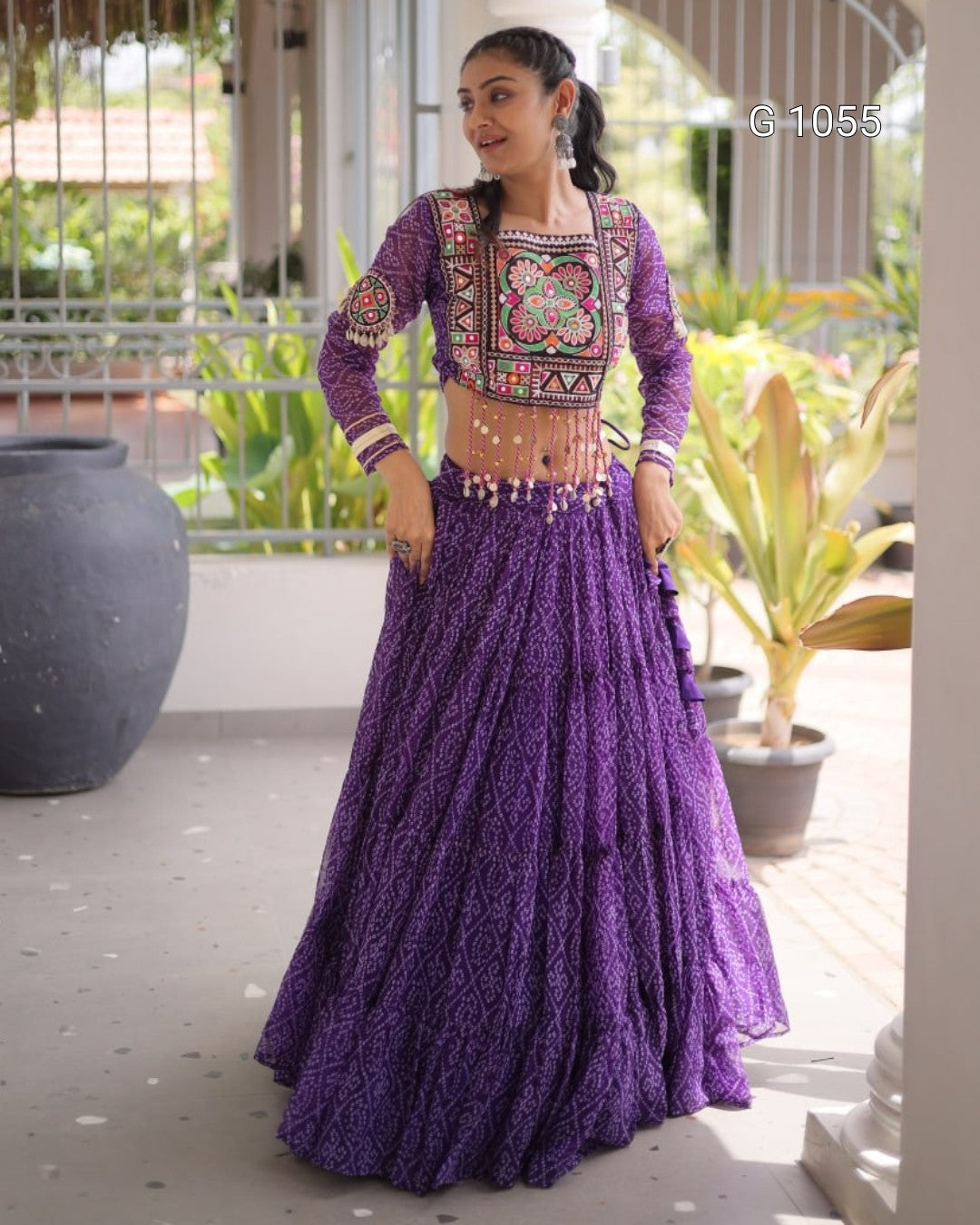 A woman modeling a purple embroidered traditional Indian outfit with a long flared skirt and patterned blouse, standing on a covered patio with plants in the background.