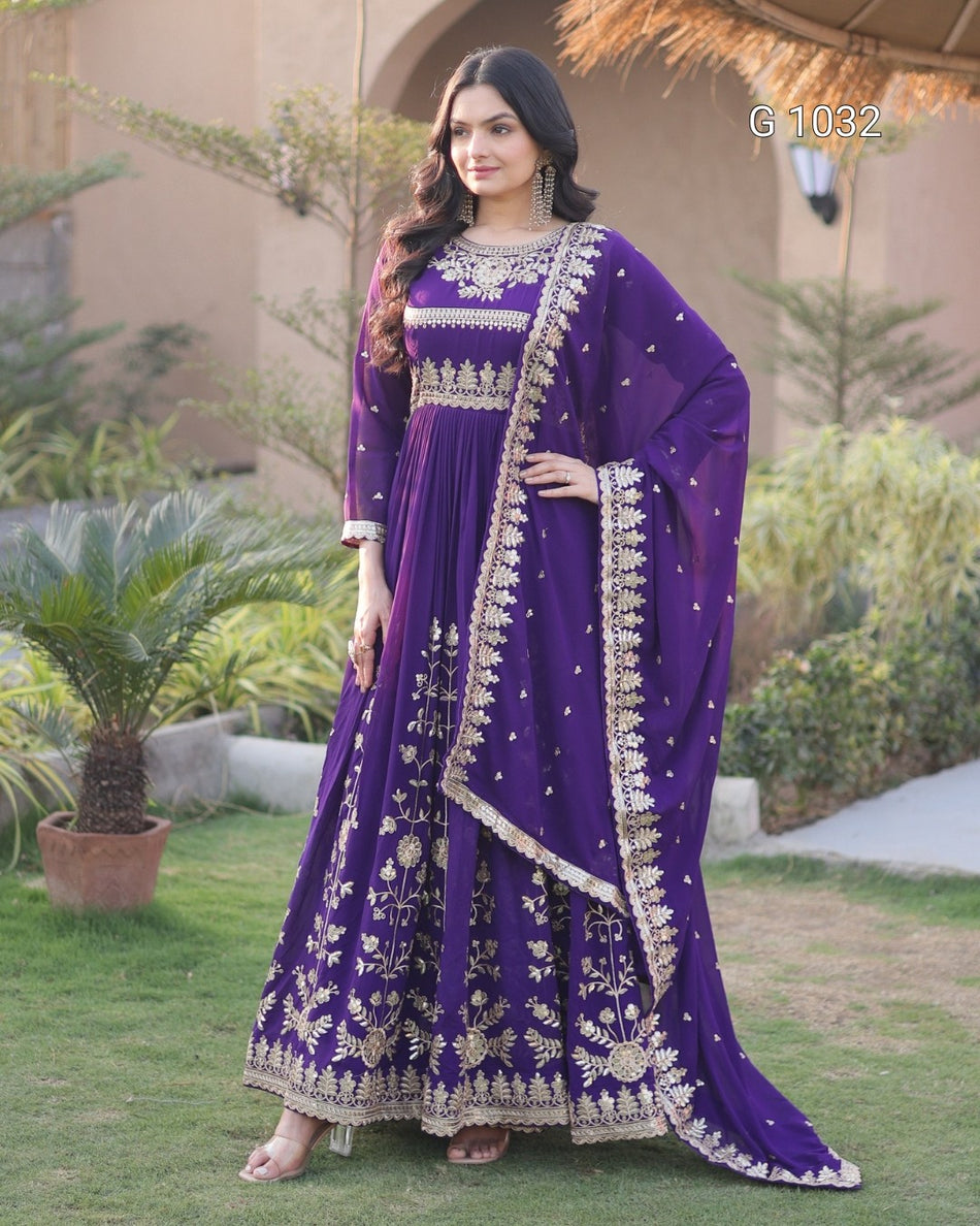 A woman stands outdoors on a lawn wearing a long purple traditional outfit with ornate gold embroidery and a matching draped scarf. The setting includes grass, plants, and a building in the background.