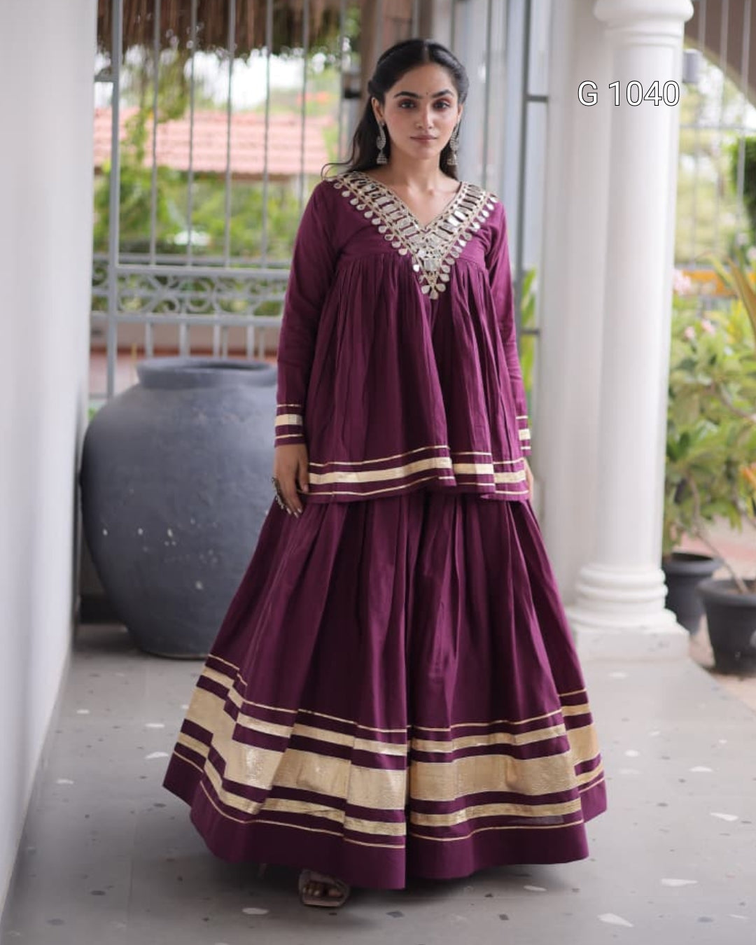 A woman modeling a deep purple traditional Indian outfit with gold zari borders and a heavily embellished silver neckline, standing on a covered porch.