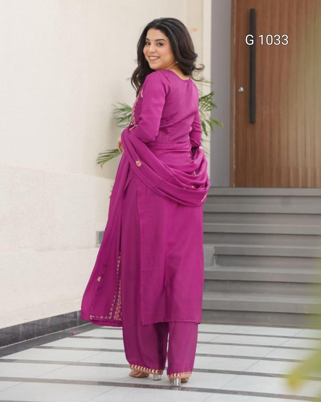 A woman modeling a magenta pink traditional three-piece salwar kameez suit with matching dupatta, standing in an indoor setting with tiled floors and a wooden door. The text 'G 1033' is visible in the top right corner.