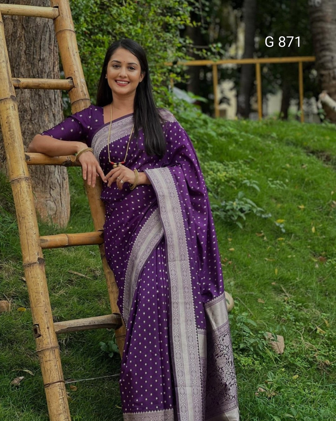 Woman in a purple saree with white patterns sitting on a wooden ladder outdoors.