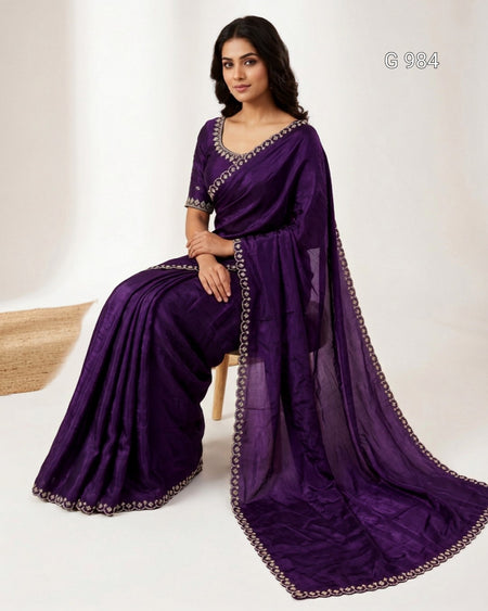 A woman seated on a small stool wearing a deep purple sari with silver embroidered borders and a matching embroidered blouse, photographed in a clean studio setting.