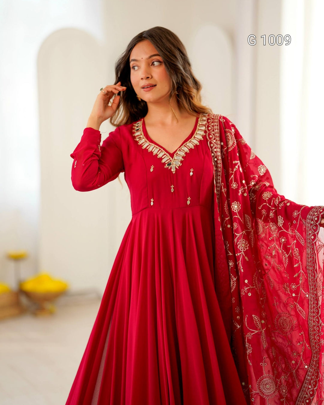 A woman modeling a long red traditional dress with gold embroidery and a matching sheer dupatta, photographed in a softly lit indoor setting with white arches in the background.