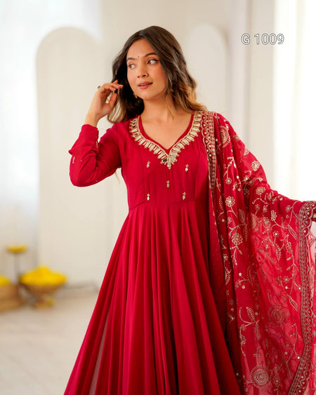 A woman modeling a long red traditional dress with gold embroidery and a matching sheer dupatta, photographed in a softly lit indoor setting with white arches in the background.