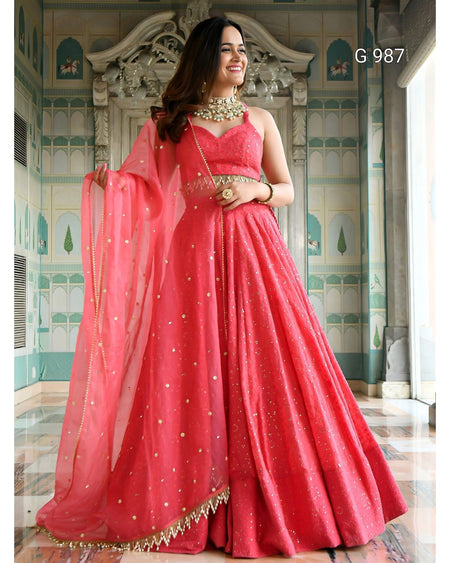 A woman models a bright pinkish-red traditional Indian lehenga choli suit with a matching dupatta, decorated with gold zari embroidery and mirror work, set against an ornate architectural background.