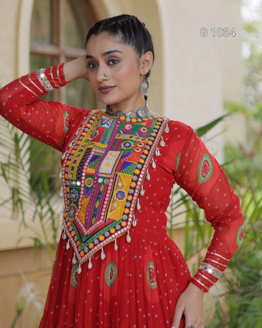 A woman wearing a red long-sleeve embroidered dress with a heavily embellished multicolor yoke panel, silver bangles, and a silver necklace, posing in front of a softly blurred beige building and green plants.