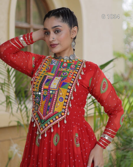 A woman wearing a red long-sleeve embroidered dress with a heavily embellished multicolor yoke panel, silver bangles, and a silver necklace, posing in front of a softly blurred beige building and green plants.