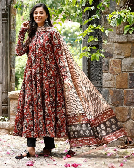 Woman in a traditional outfit with a patterned shawl standing outdoors near stone walls and greenery.