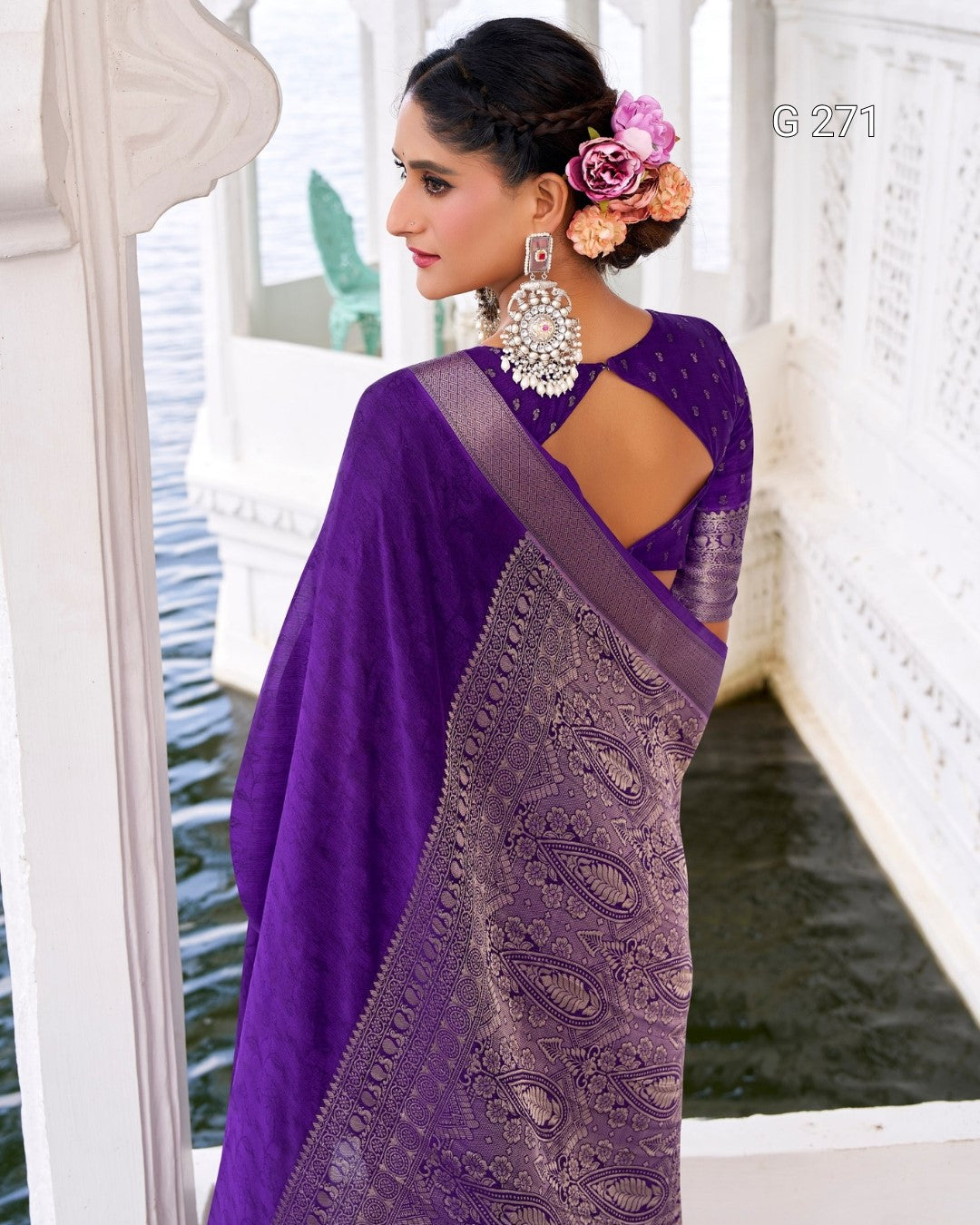 A woman wearing a purple saree with a matching embroidered blouse, standing in a white architectural setting near water. The saree has a sheer purple body with a metallic lavender paisley border, and she is wearing large ornate earrings.