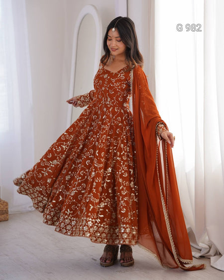 A woman modeling a rust-orange embroidered Anarkali gown with a matching dupatta, standing in a bright white room with sheer curtains. The dress features intricate gold and silver floral embroidery and mirror work, and the model is holding out the flared skirt to show its volume.
