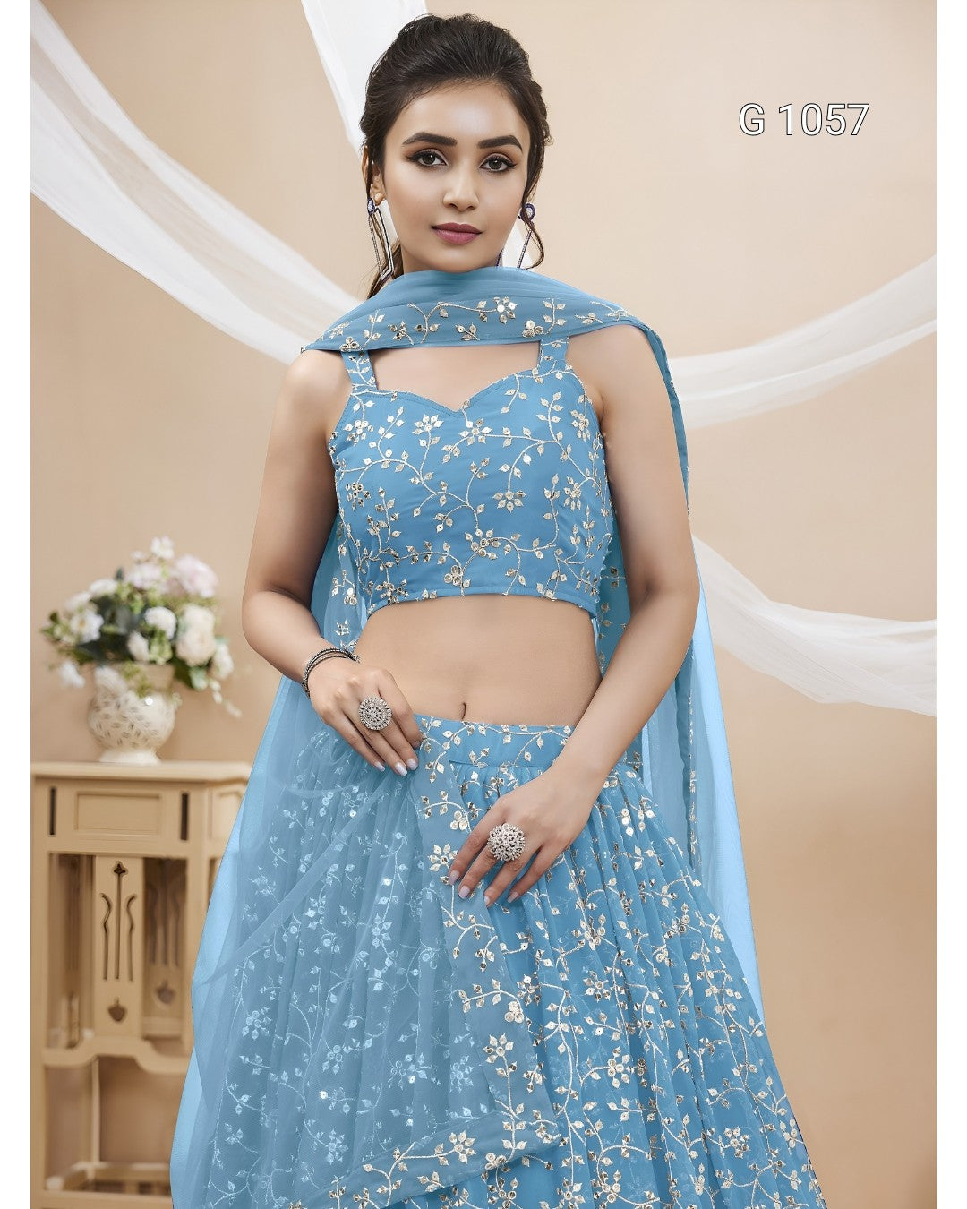 A woman modeling a light blue embroidered Indian ethnic outfit with a sleeveless crop top, flared skirt, and matching dupatta. The outfit has white floral embroidery, and the background is a beige studio setting with decorative drapes and a small table with flowers.