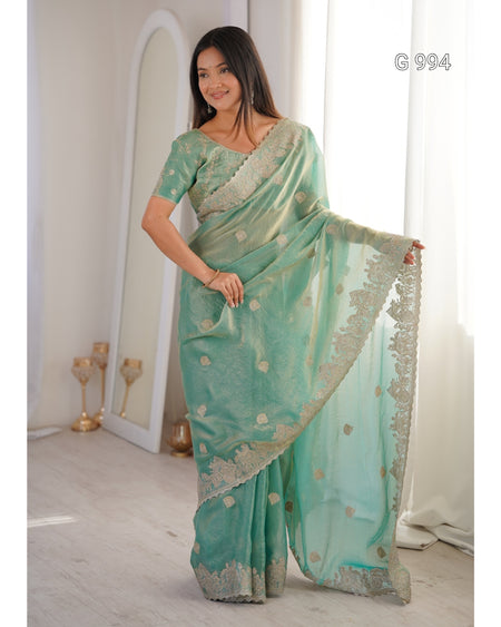 A woman modeling a mint green saree with gold floral motifs and a gold embroidered border, standing in a bright indoor setting with a mirror and curtains in the background.