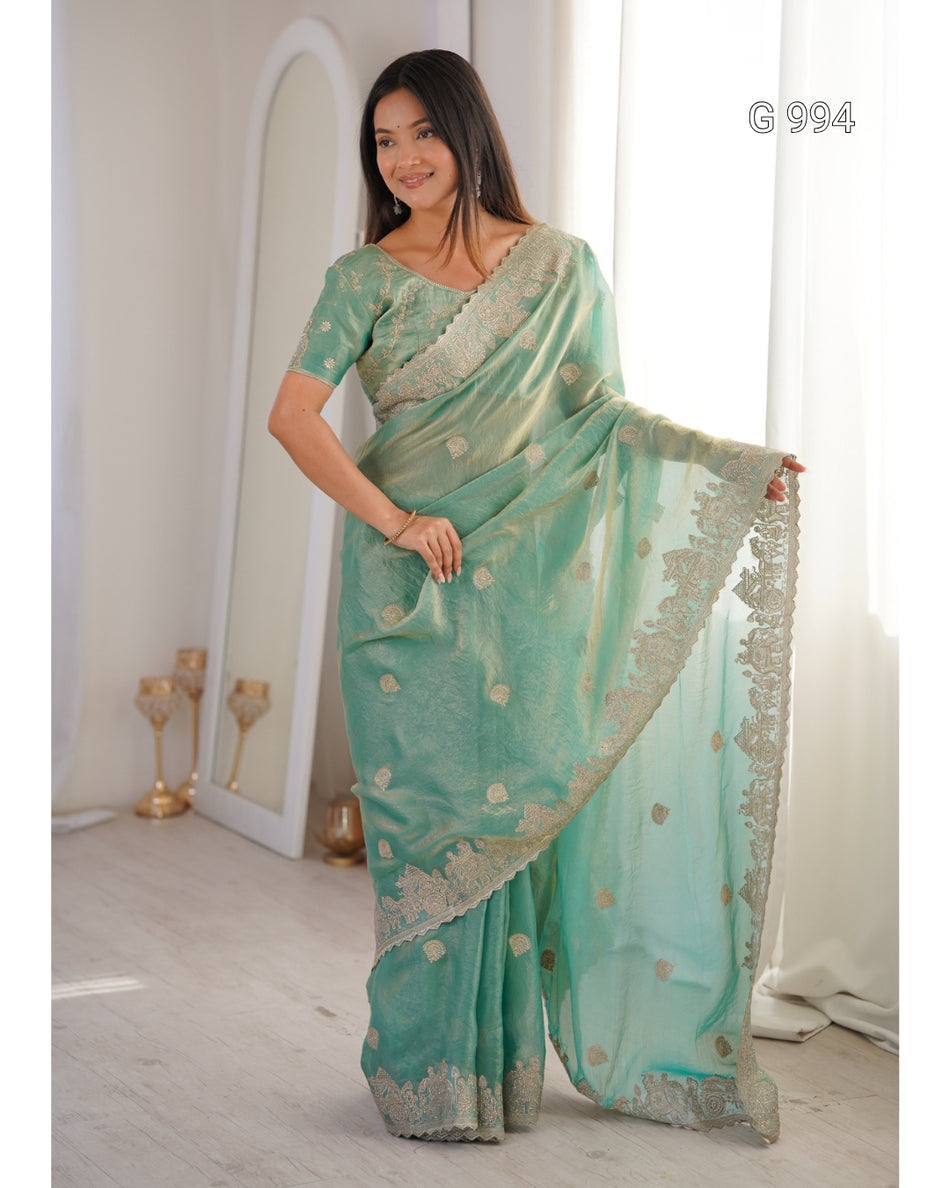 A woman modeling a mint green saree with gold floral motifs and a gold embroidered border, standing in a bright indoor setting with a mirror and curtains in the background.
