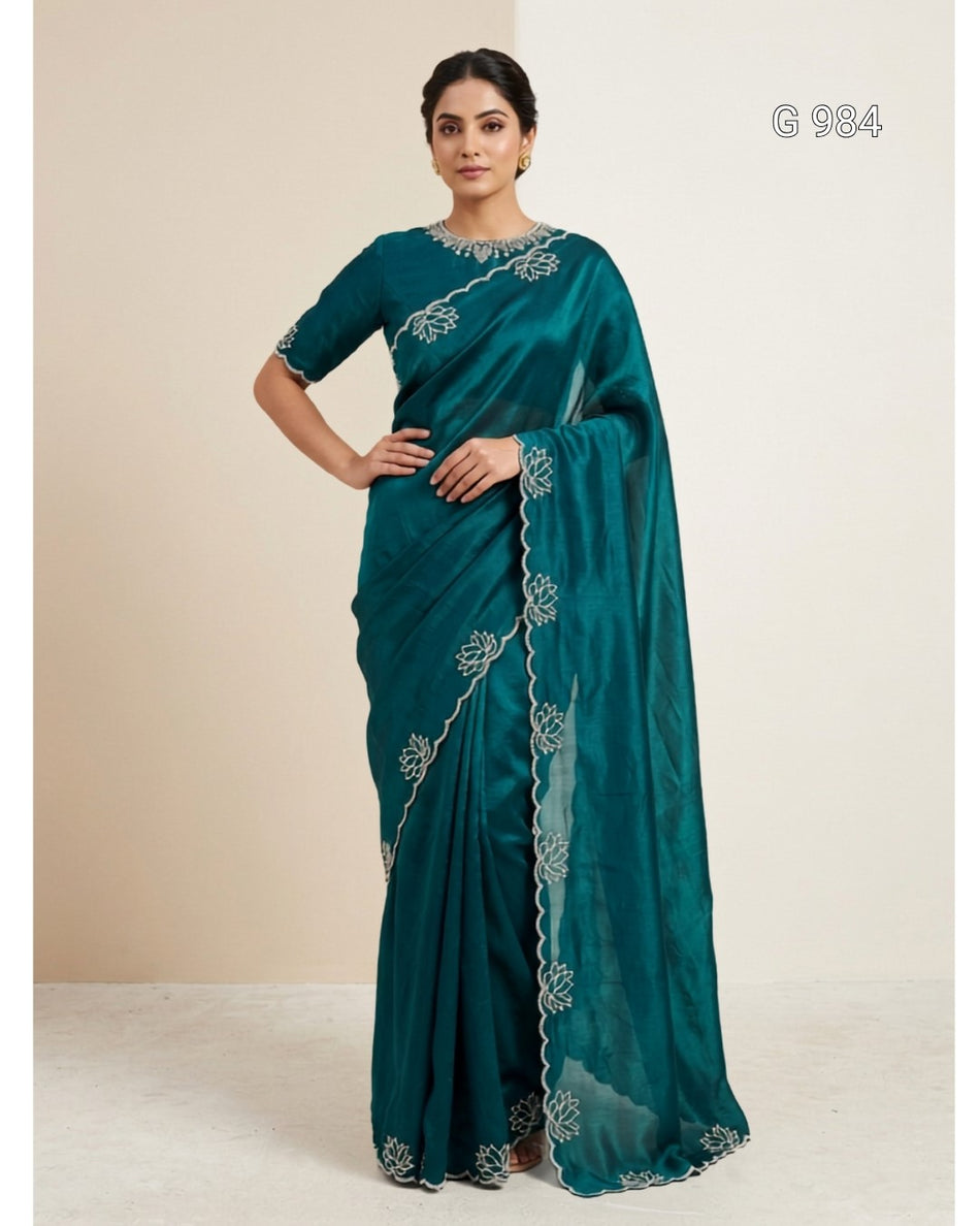 A woman modeling a teal green saree with silver floral embroidery and a matching embroidered border, standing against a plain beige studio background.