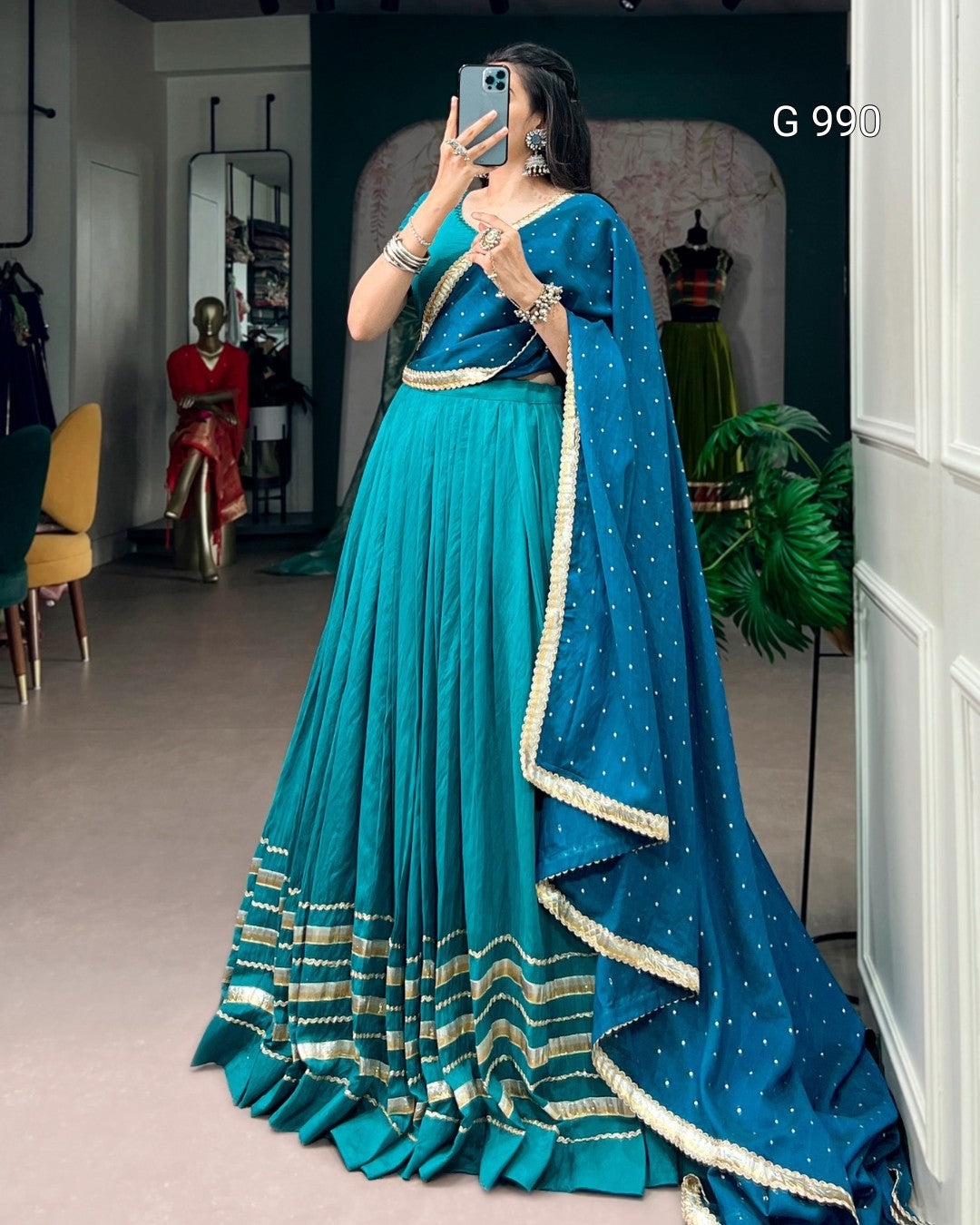 A woman takes a mirror selfie in a boutique wearing a traditional Indian outfit consisting of a teal blue skirt (lehenga), a matching blouse, and a draped dupatta with gold zari borders and star patterns. The background shows other clothing items on mannequins and store fixtures.