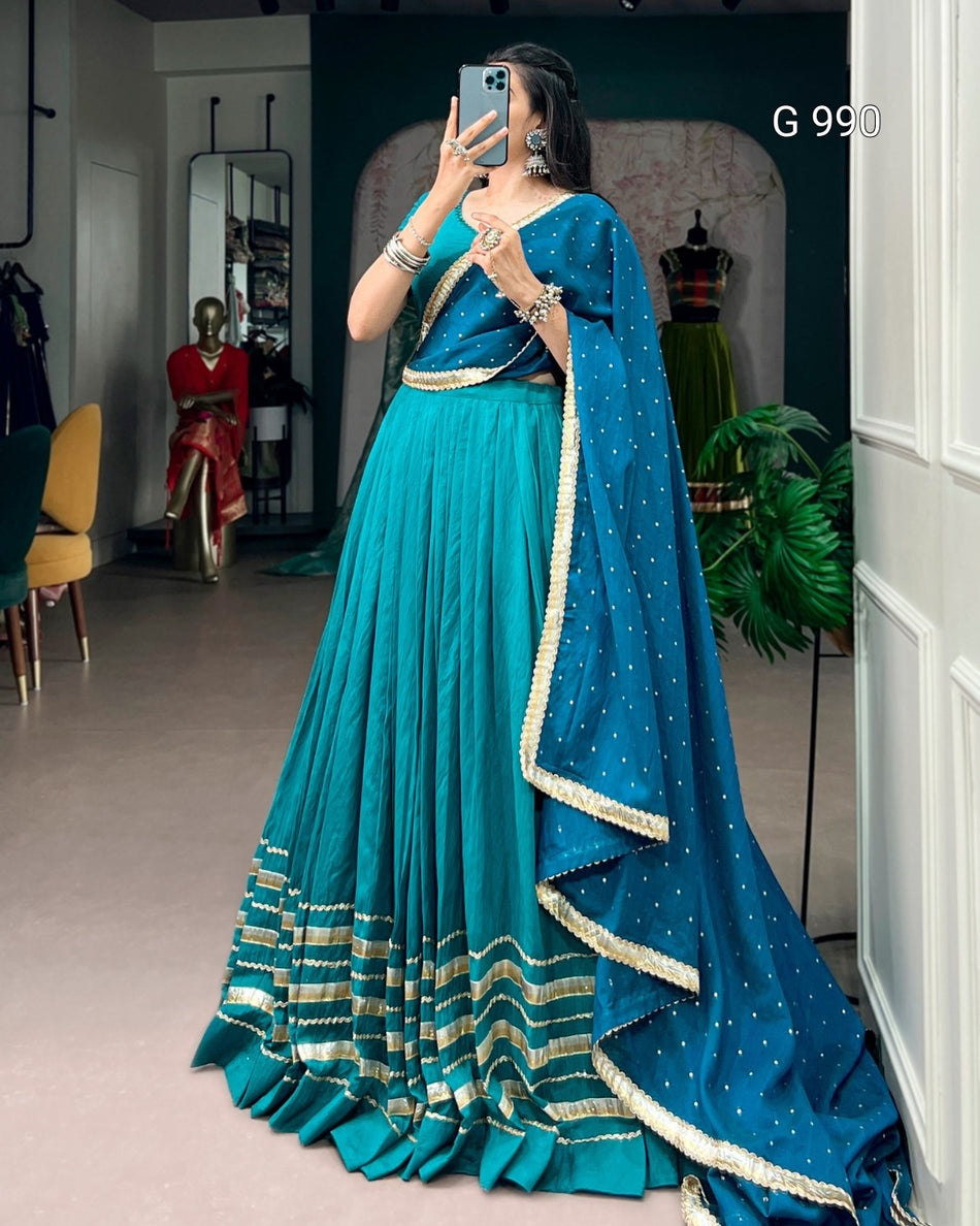 A woman takes a mirror selfie in a boutique wearing a traditional Indian outfit consisting of a teal blue skirt (lehenga), a matching blouse, and a draped dupatta with gold zari borders and star patterns. The background shows other clothing items on mannequins and store fixtures.