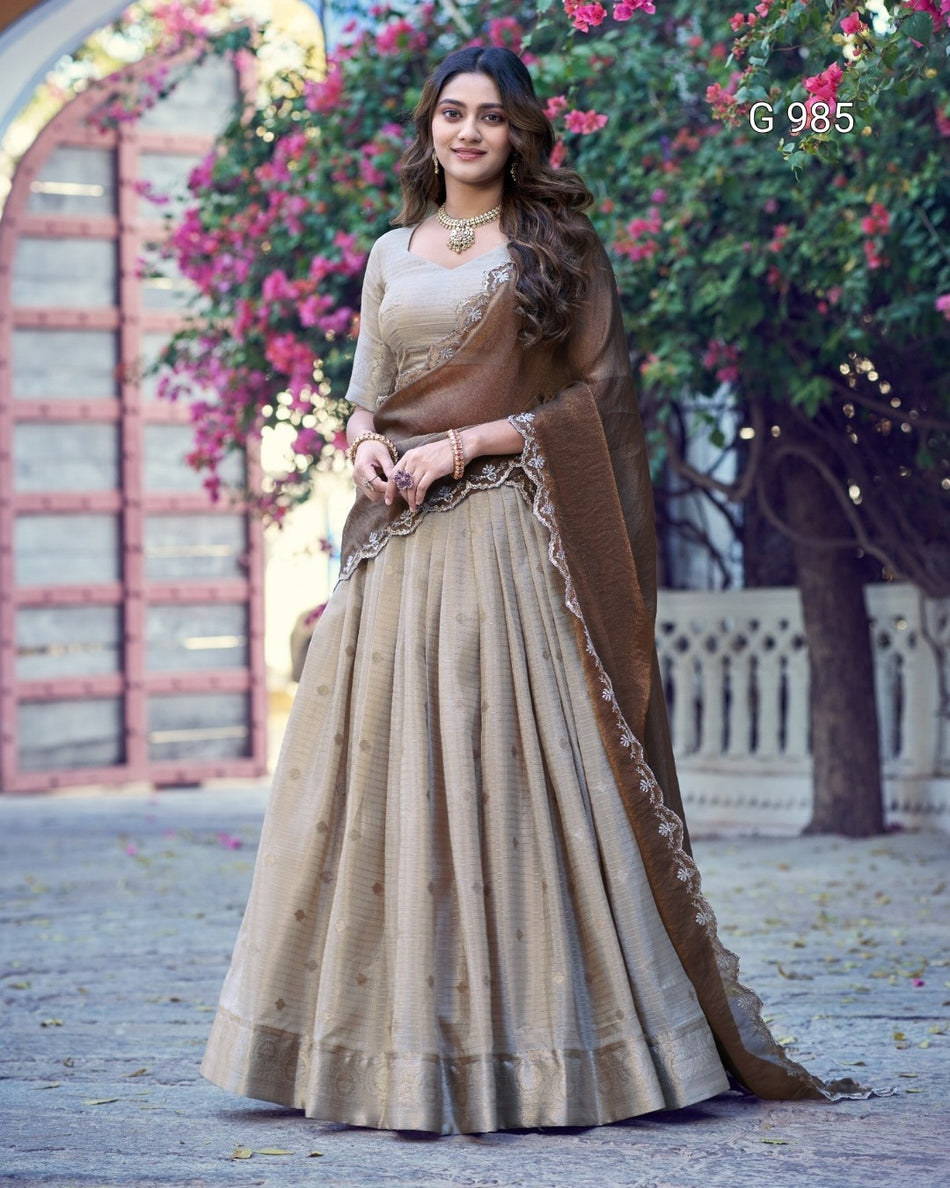 A woman wearing a beige and brown traditional Indian outfit with a long flared skirt, embroidered blouse, and draped dupatta, standing outdoors in front of flowering trees and a stone structure. The text 'G 985' appears in the top right.