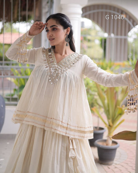 A woman modeling a cream-colored traditional outfit with intricate silver embroidery and gold accents, set against a blurred outdoor patio background with potted plants.