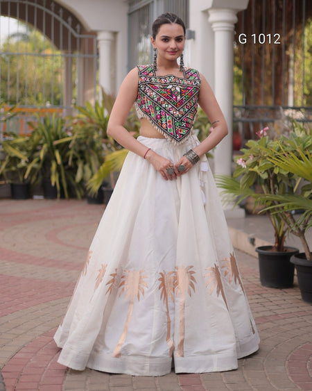 A woman modeling a traditional Indian outfit consisting of a sleeveless white skirt with gold palm tree prints and a heavily embroidered multicolor crop top, standing in an outdoor courtyard.