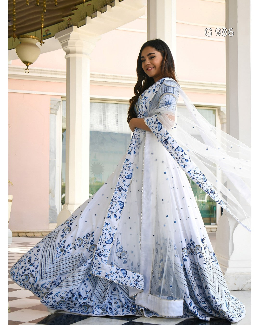 A woman modeling a white traditional dress with blue floral and geometric patterns, standing on a porch with columns. The dress features a flared skirt, sheer sleeves, and a matching dupatta.