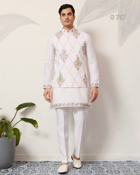 Man wearing a white embroidered kurta with white pants standing against a light gray wall.