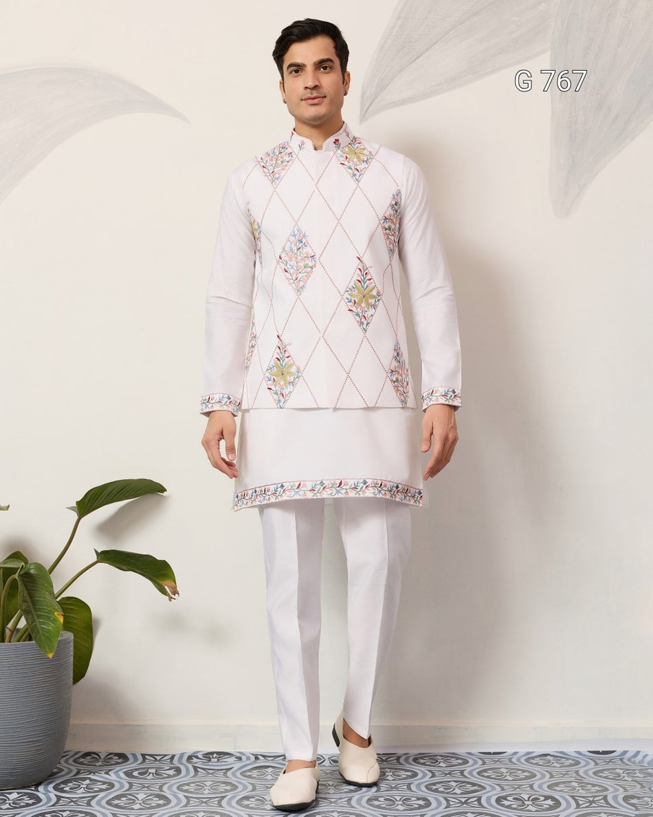 Man wearing a white embroidered kurta with white pants standing against a light gray wall.