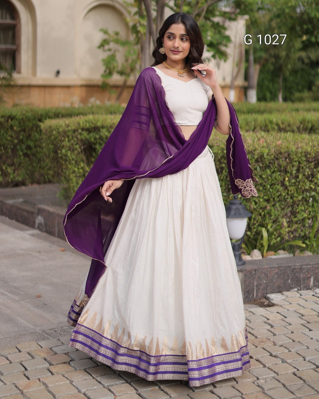 A woman models a traditional Indian outfit featuring an off-white/cream lehenga skirt with a purple and gold border, a matching off-white blouse, and a sheer purple dupatta draped over her shoulders. The background shows a garden path and a building.