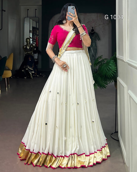 A woman modeling a traditional Indian outfit consisting of a white flared lehenga skirt with gold zari borders and small metallic embellishments, paired with a bright pink blouse and a gold and pink draped scarf. She is taking a mirror selfie in an indoor showroom setting.