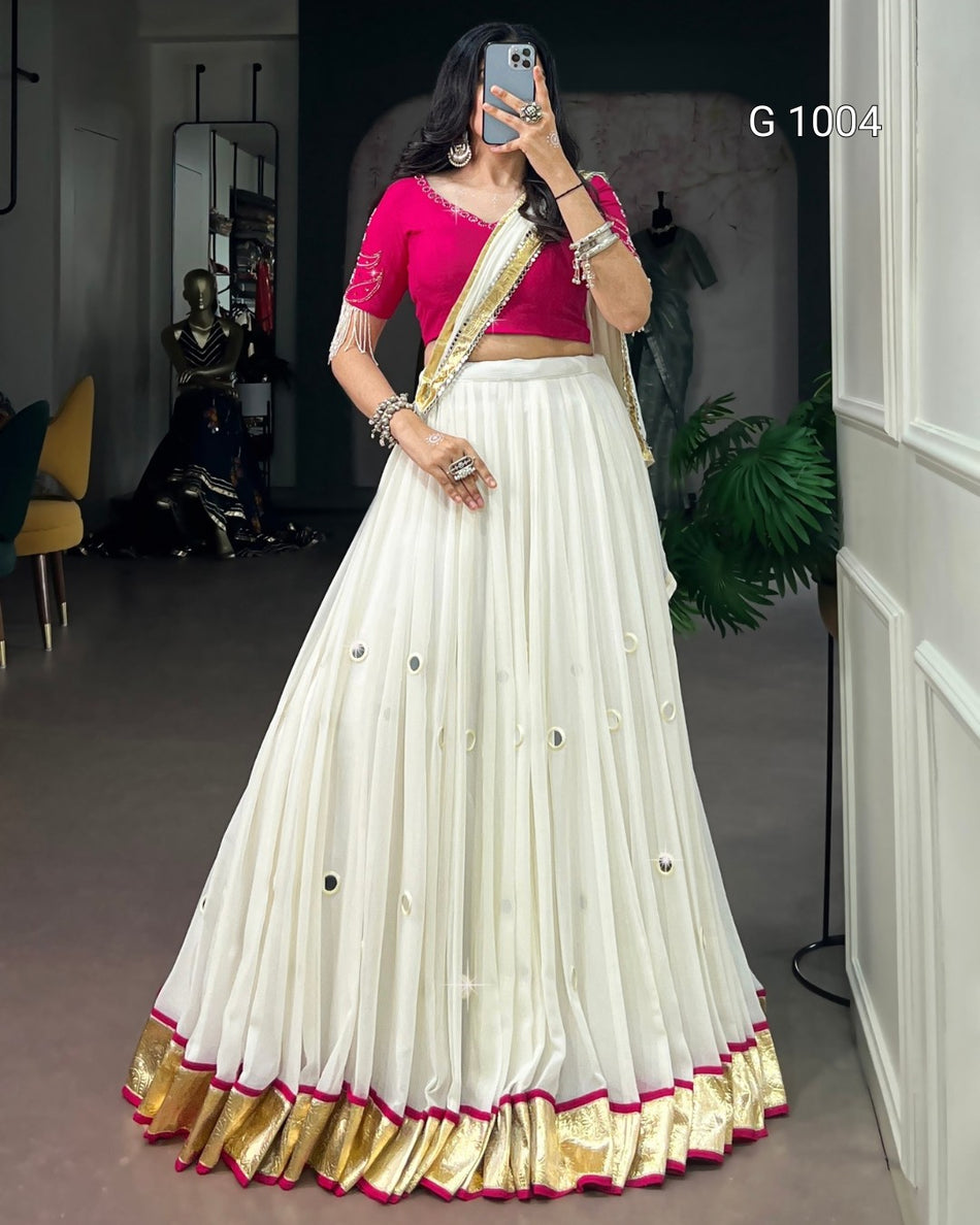 A woman modeling a traditional Indian outfit consisting of a white flared lehenga skirt with gold zari borders and small metallic embellishments, paired with a bright pink blouse and a gold and pink draped scarf. She is taking a mirror selfie in an indoor showroom setting.