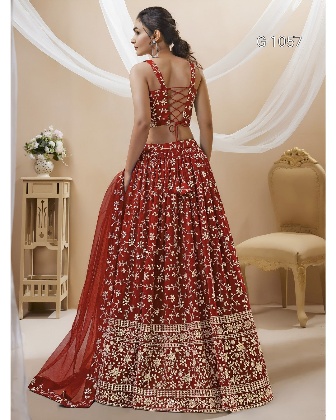 A woman modeling a red and gold embroidered lehenga choli set, shown from the back in a decorated studio setting with draped white fabric, a side table with flowers, and a gold chair.