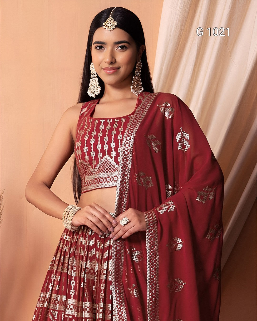 A woman modeling a traditional Indian outfit consisting of a maroon red and gold patterned sleeveless crop top, matching pleated skirt, and a draped dupatta with gold floral motifs, set against a warm beige background.