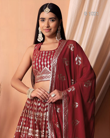 A woman modeling a traditional Indian outfit consisting of a maroon red and gold patterned sleeveless crop top, matching pleated skirt, and a draped dupatta with gold floral motifs, set against a warm beige background.