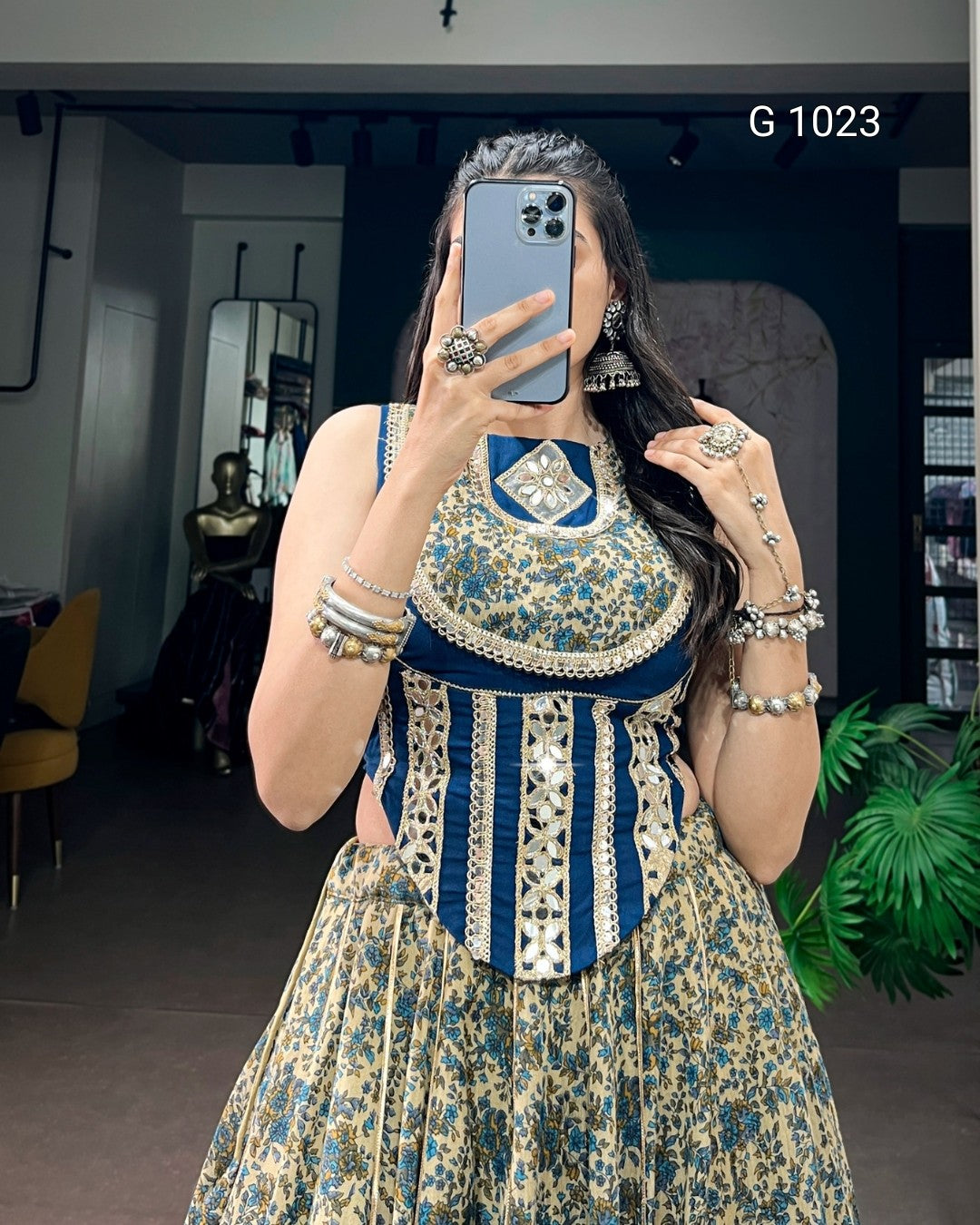 A woman takes a mirror selfie in a boutique wearing a sleeveless, heavily embellished traditional dress with a blue and beige floral pattern, a solid navy blue collar, and vertical silver beadwork on the bodice. She is accessorized with silver jewelry, including bracelets, rings, and earrings.