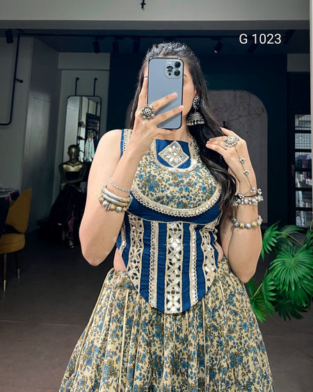 A woman takes a mirror selfie in a boutique wearing a sleeveless, heavily embellished traditional dress with a blue and beige floral pattern, a solid navy blue collar, and vertical silver beadwork on the bodice. She is accessorized with silver jewelry, including bracelets, rings, and earrings.