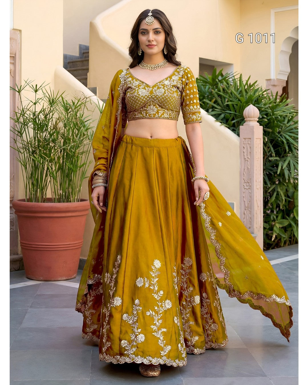 A woman modeling a mustard yellow and gold embroidered Indian ethnic wear set, featuring a flared skirt with floral embroidery, a matching embroidered blouse, and a sheer dupatta, set against an outdoor architectural background.
