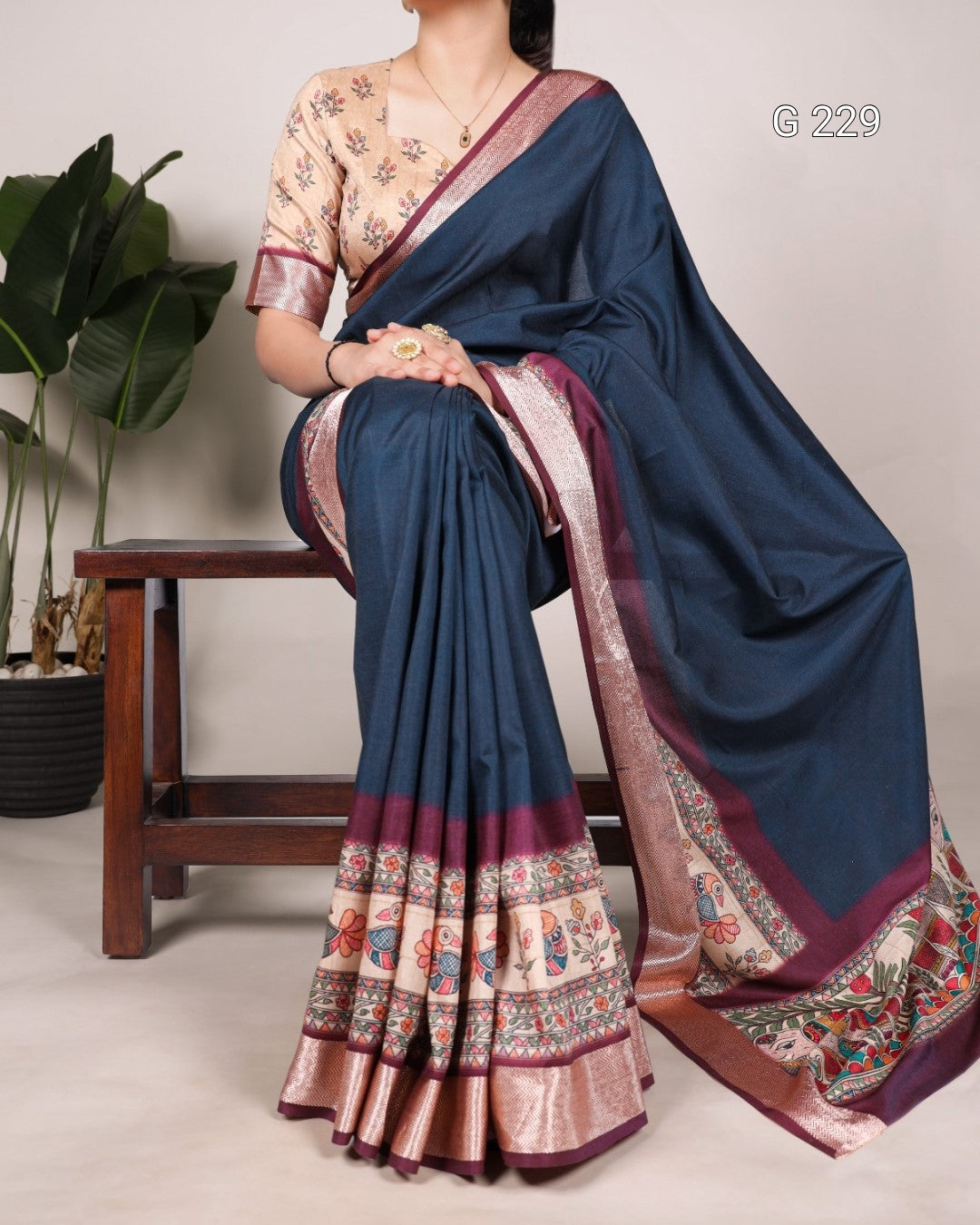 Woman wearing a blue saree with a patterned border, sitting on a wooden stool.