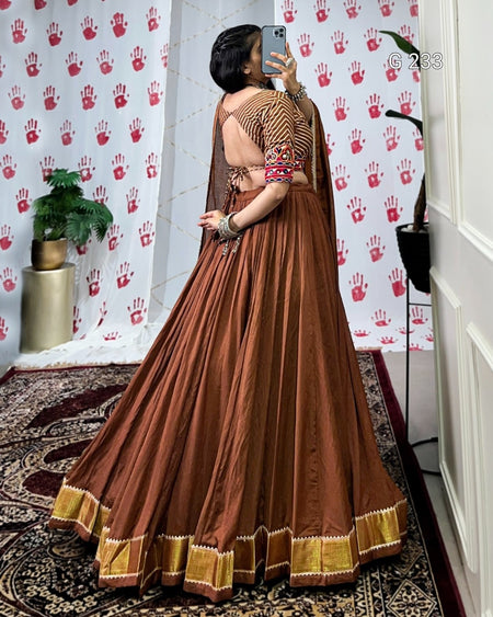Woman in a brown saree with gold border taking a mirror selfie against a decorative wall.