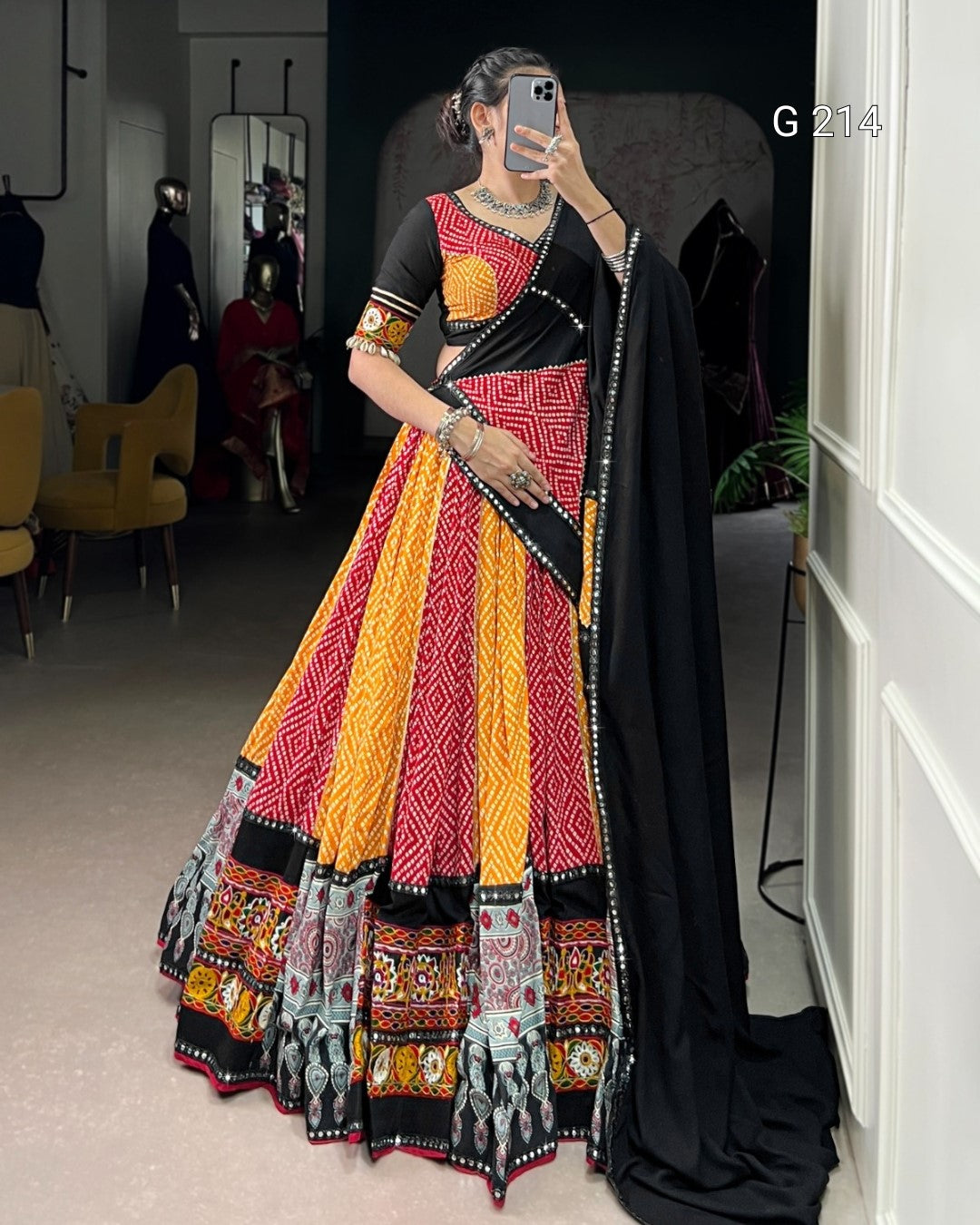 Person wearing a colorful traditional outfit with a black dupatta in a room.