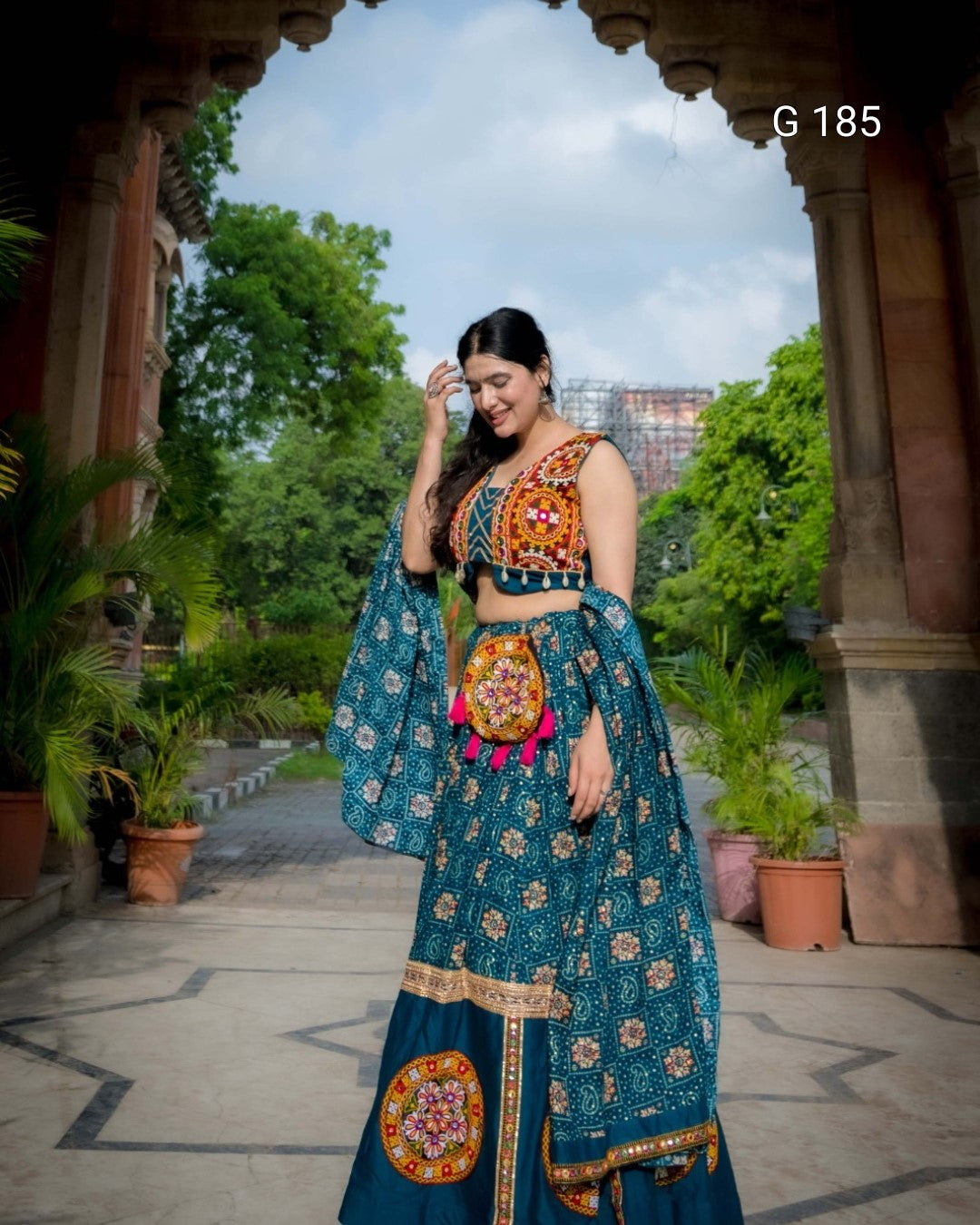 Woman in traditional outfit standing outdoors with greenery and architecture in the background