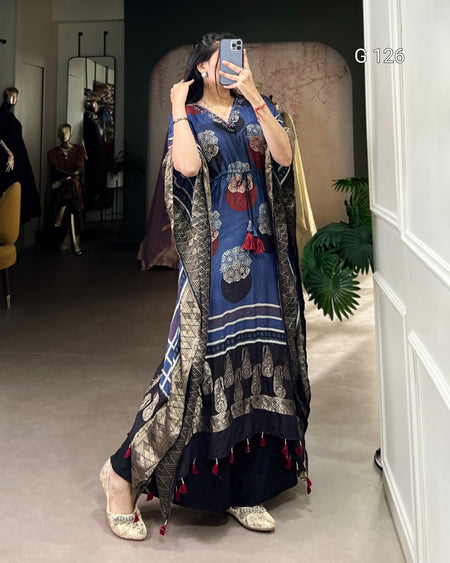 Woman taking a mirror selfie wearing a long, patterned dress in a room with a plant and chair.