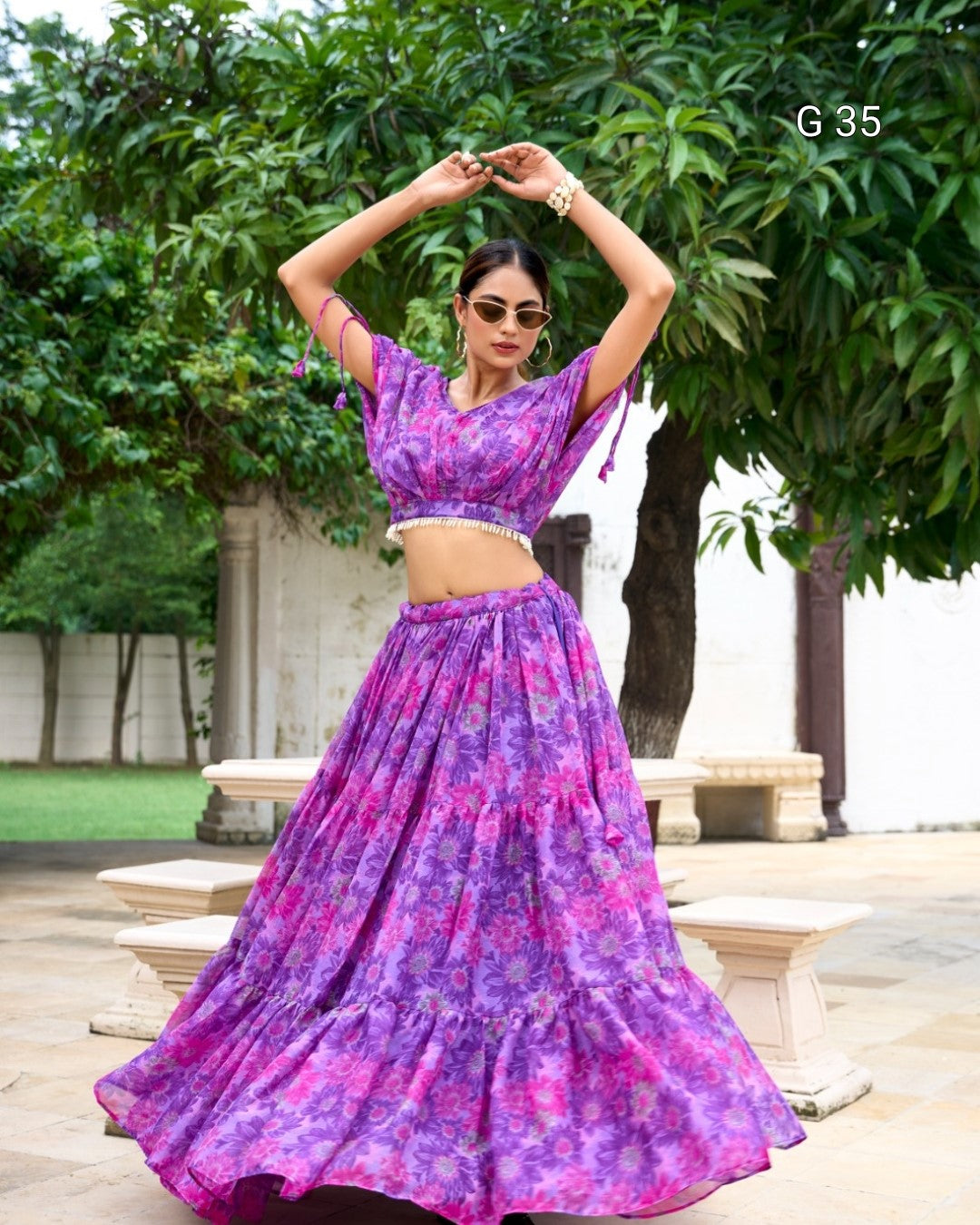 Woman in a purple floral outfit standing outdoors with greenery in the background