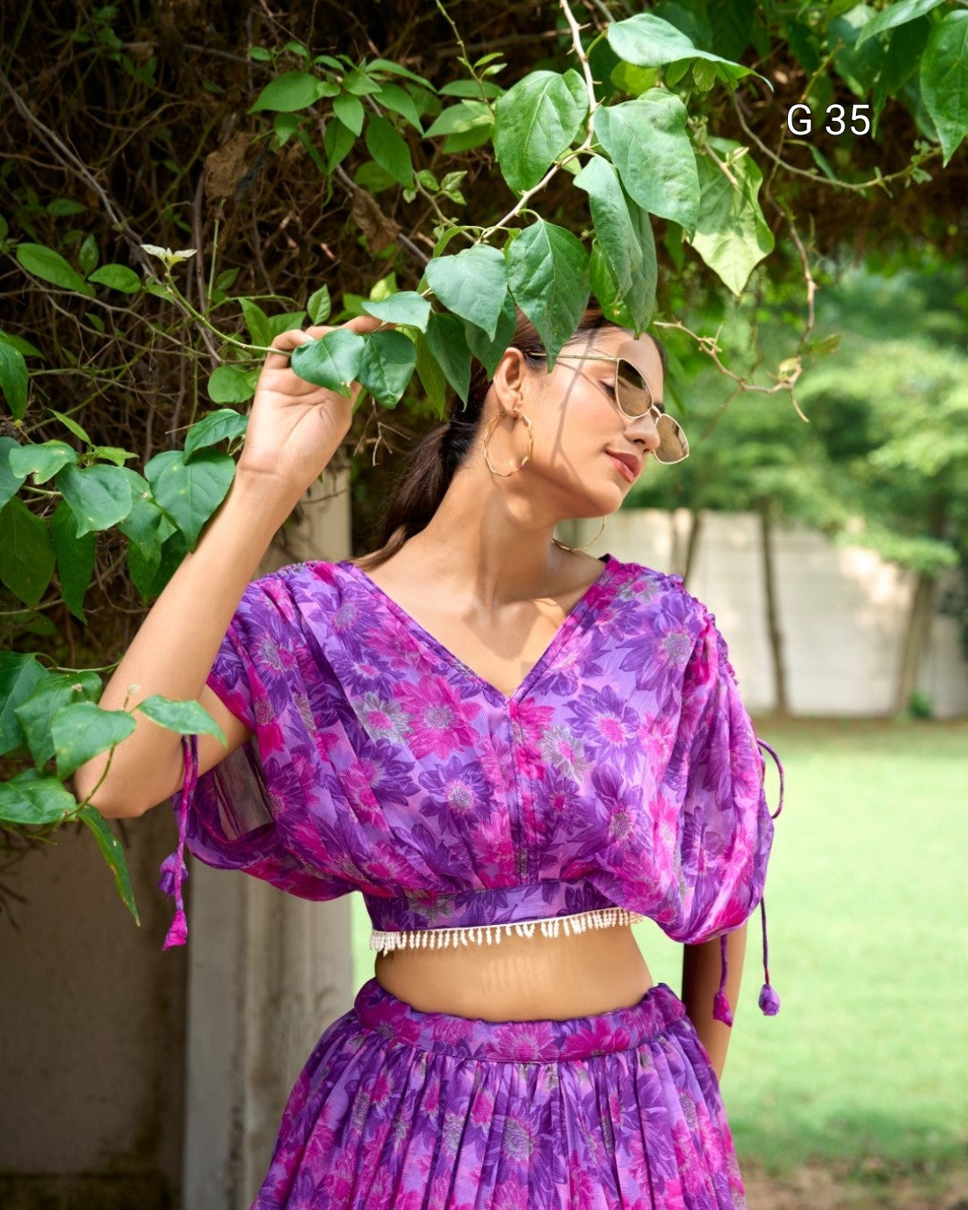 Woman wearing a purple floral outfit standing outdoors with greenery around
