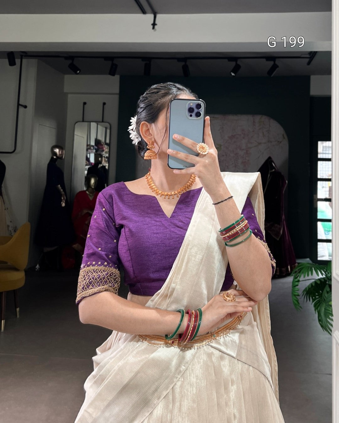 Woman taking a mirror selfie wearing a purple blouse and white saree with jewelry.
